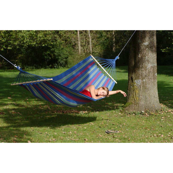 Person relaxing in a colorful striped hammock under a tree in a park.
