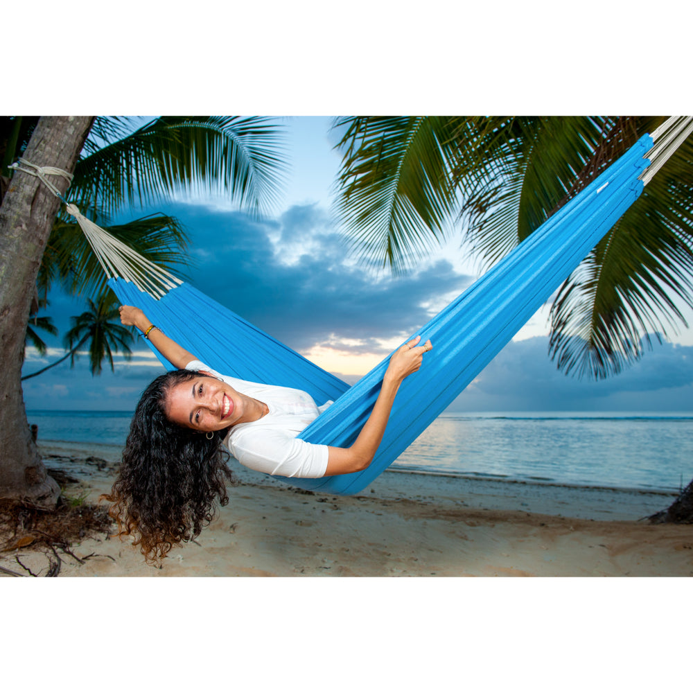 Woman relaxing in a blue hammock on a tropical beach with palm trees and ocean view.