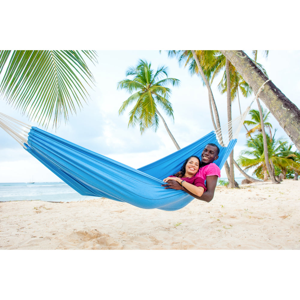 Two people in a blue hammock on a tropical beach with palm trees.
