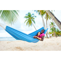 Two people in a blue hammock on a tropical beach with palm trees.