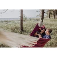 Two people in a red hammock surrounded by nature with trees and grass.