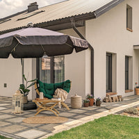 An outdoor patio setup in front of a modern white house, featuring a large dark patio umbrella, a green tufted lounge chair with a beige throw blanket, a small wooden side table, potted plants, and a stone-tiled floor.