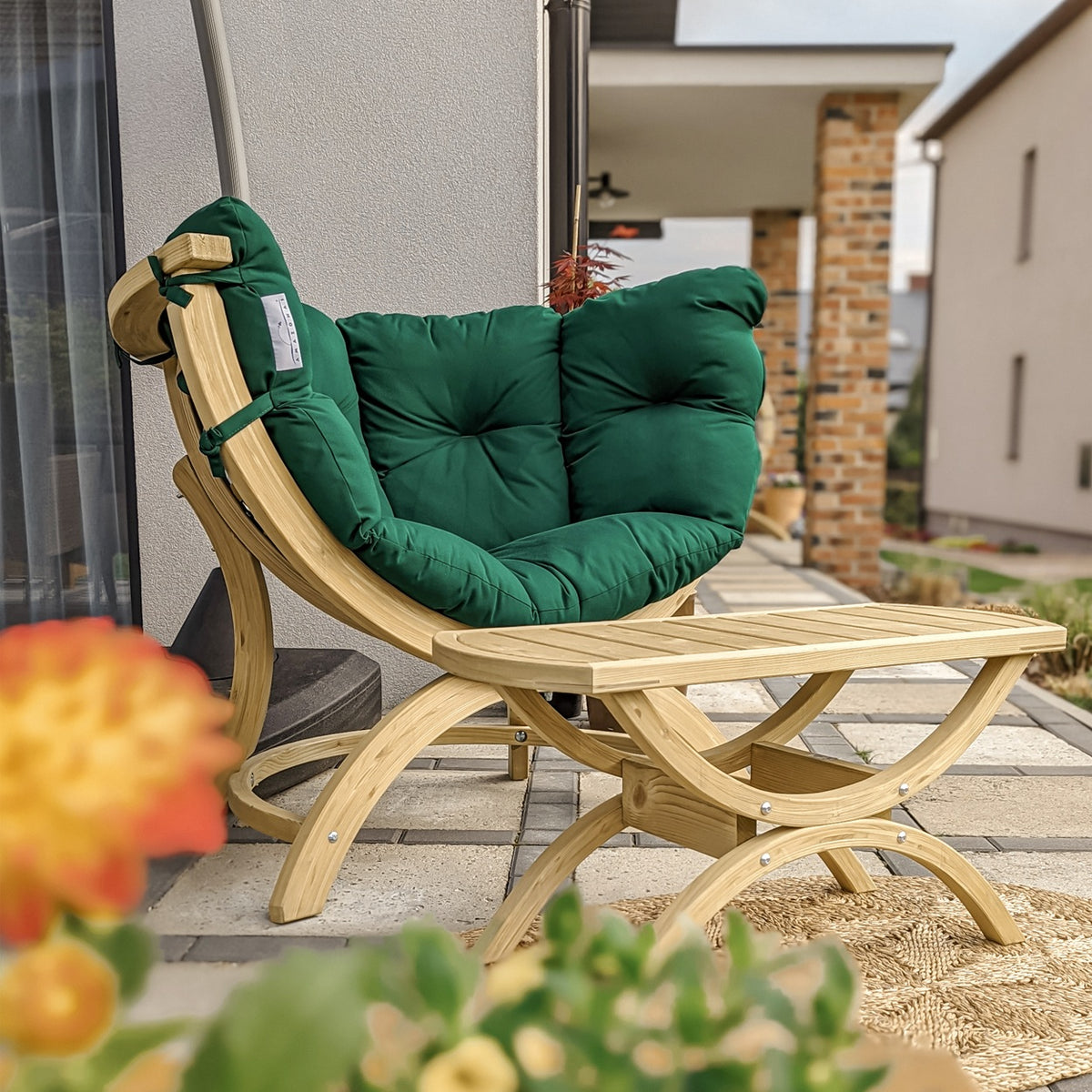A wooden outdoor lounge chair with green tufted cushions, placed on a patio in front of a house with a blurred flower in the foreground.