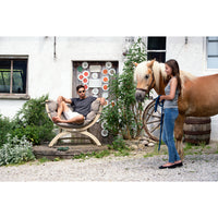 A man relaxes in a large cushioned chair set outdoors next to a woman holding a brown horse. The setting features a rustic white building, a decorative wooden door, a vintage wagon wheel, and gravel ground.