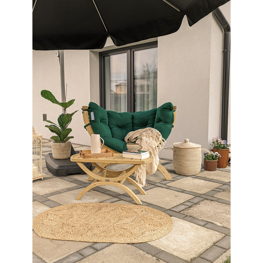 A dark green tufted outdoor lounge chair with a light wood frame sits on a stone patio under a black patio umbrella, styled with a small wooden side table, a beige throw blanket, a potted plant, and woven baskets.