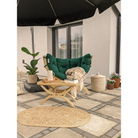 A dark green tufted outdoor lounge chair with a light wood frame sits on a stone patio under a black patio umbrella, styled with a small wooden side table, a beige throw blanket, a potted plant, and woven baskets.