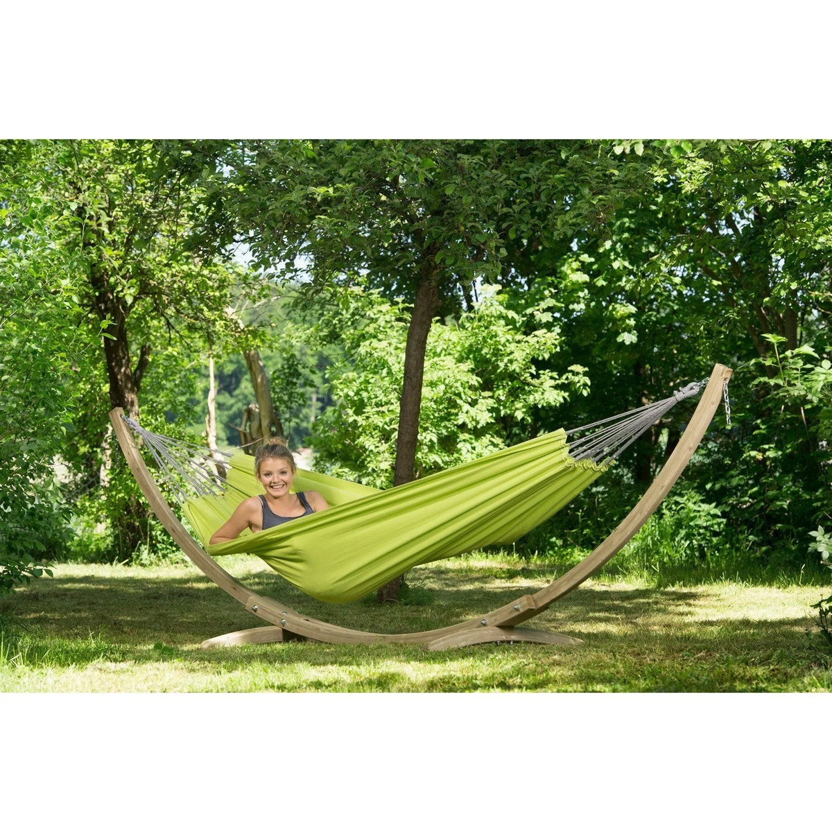 Person relaxing in a green hammock with a wooden stand in a park setting.