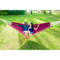 Two people relaxing in a burgundy hammock on a grassy field.