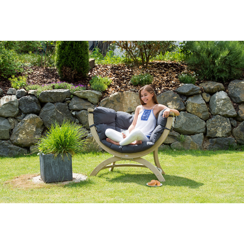 A woman sits in a curved outdoor lounge chair with dark gray cushions on a grassy lawn, with a stone wall and garden plants in the background.