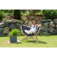 A woman sits in a curved outdoor lounge chair with dark gray cushions on a grassy lawn, with a stone wall and garden plants in the background.