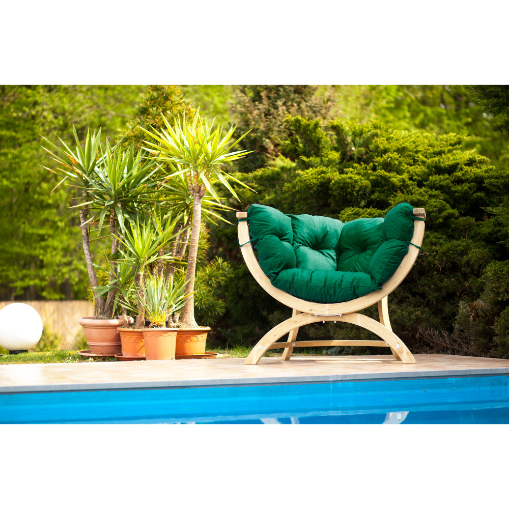 A curved outdoor lounge chair with a natural wood frame and plush green cushions, positioned beside a swimming pool with potted plants and green shrubs in the background.