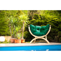 A curved outdoor lounge chair with a natural wood frame and plush green cushions, positioned beside a swimming pool with potted plants and green shrubs in the background.
