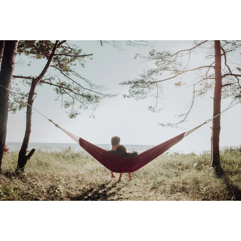 Person relaxing in a red hammock between two trees with a scenic background.