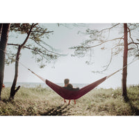 Person relaxing in a red hammock between two trees with a scenic background.
