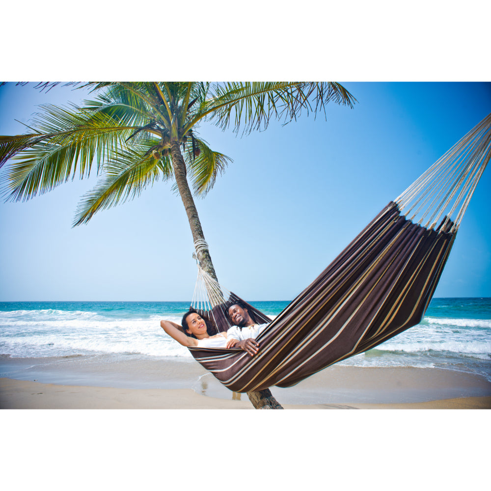 Two people relaxing in a striped hammock on a tropical beach with palm trees and blue sky.