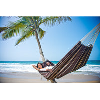 Two people relaxing in a striped hammock on a tropical beach with palm trees and blue sky.