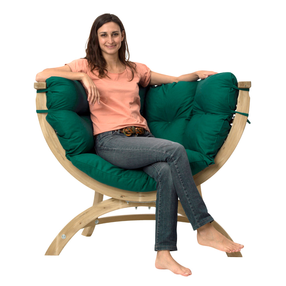 A woman sits barefoot on a curved wooden lounge chair with thick dark green cushions