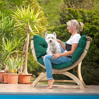 A woman sits barefoot on a curved wooden outdoor lounge chair with thick green cushions, holding a small white dog in a garden next to a pool.